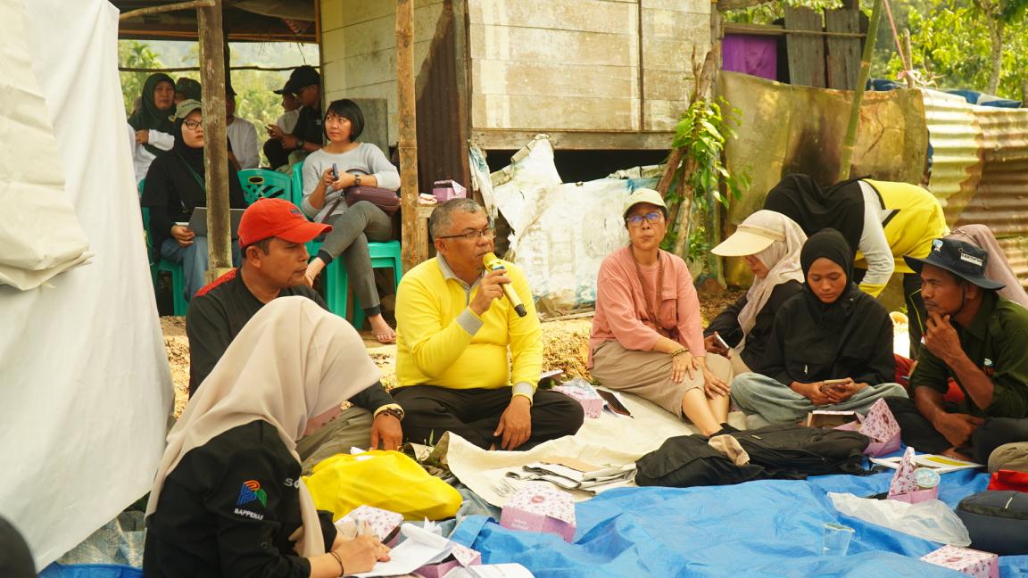 Photograph showing a group of people gathered outdoors, sitting on mats and tarps under a rustic shelter. The scene highlights a man in a yellow shirt speaking into a microphone, suggesting a community meeting or discussion, with others attentively listening or engaged in activities.