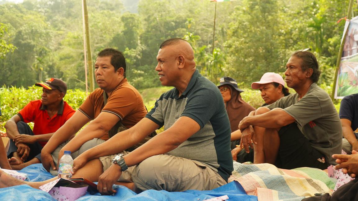 Photograph of a group of people sitting on blankets outdoors in a natural, green setting, appearing engaged in a discussion or meeting.