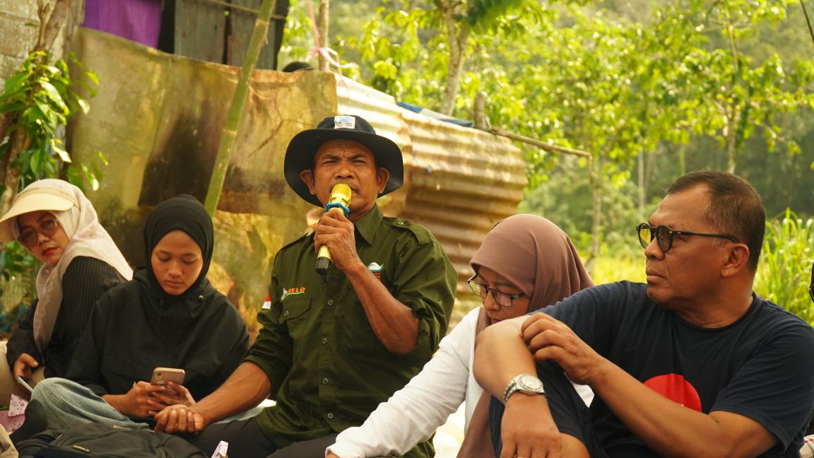 Photograph of a group of five people sitting outdoors in a natural, green setting with trees and plants in the background. A man in a green shirt and hat is speaking into a microphone, while others listen attentively.