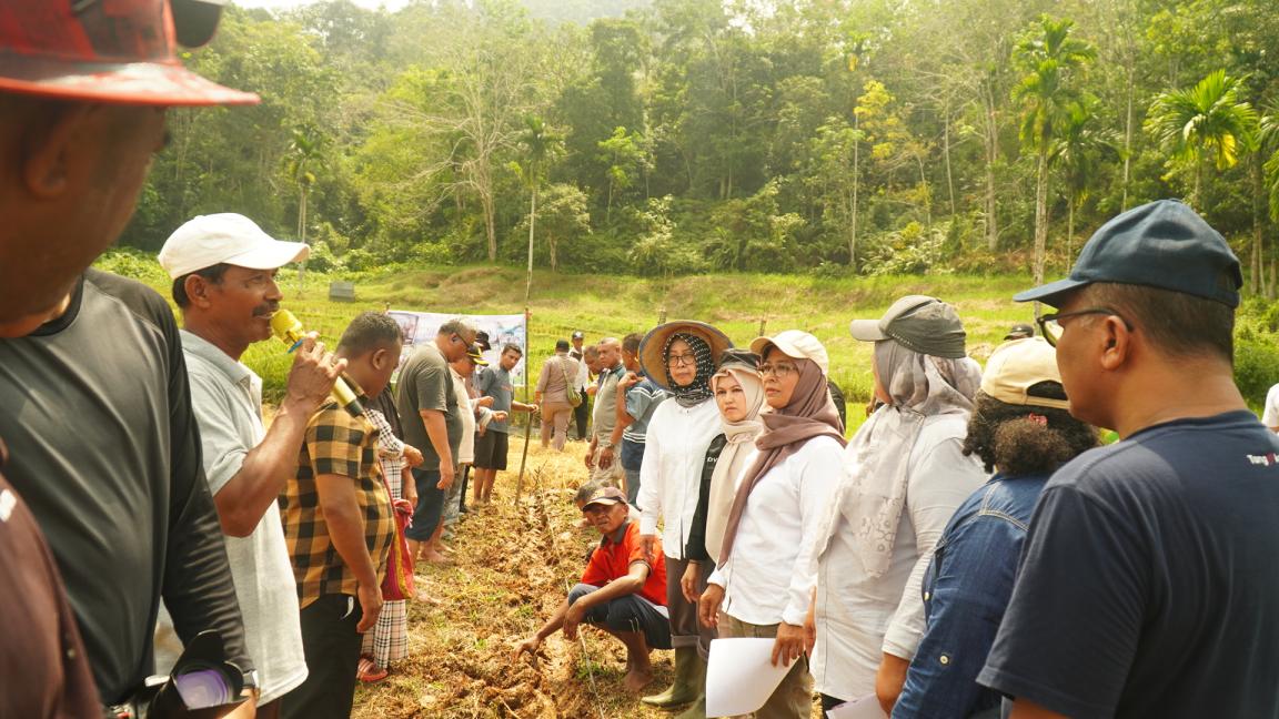 Photograph of a group of people gathered outdoors in a rural, forested area, seemingly engaged in a discussion or demonstration related to agriculture or land use. Individuals wear casual clothing and hats, with some holding papers, and a trench or dug-out section of soil runs through the center, indicating a focus on farming or environmental practices.