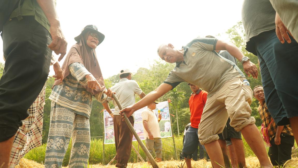 Photograph showing a group of people outdoors, with one man bending forward to hold a walking stick offered by a woman wearing a hat and patterned clothing.
