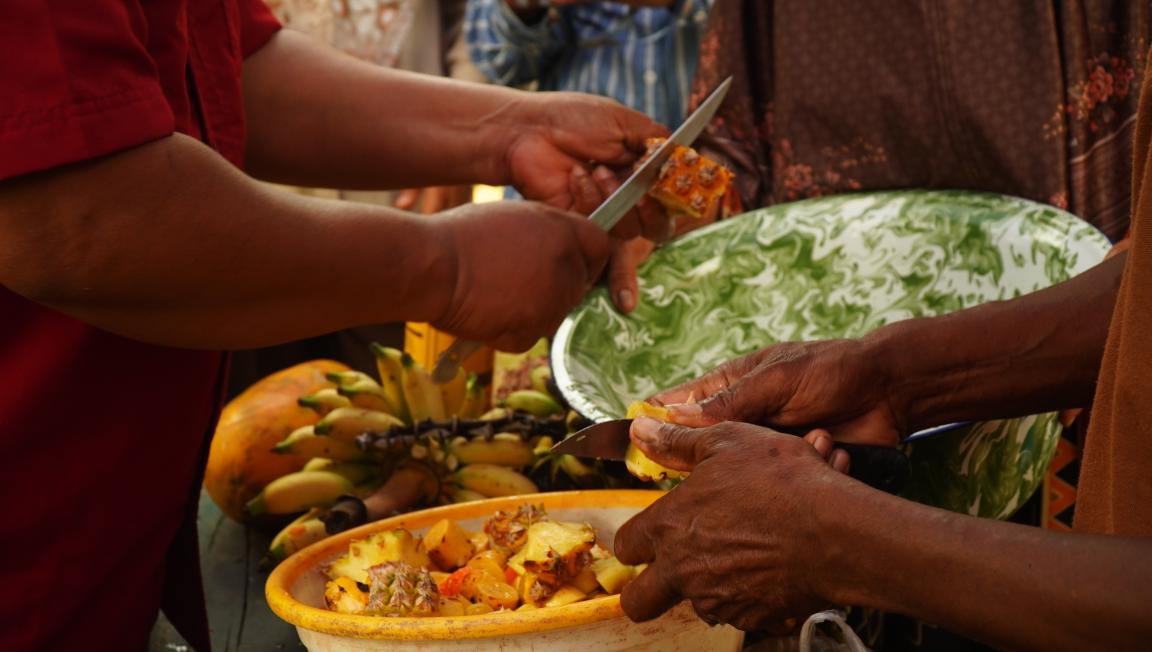 Photograph showing close-up of hands cutting and serving fresh pineapple pieces into a green and white marbled bowl. Background includes bunches of bananas and other fruits, highlighting a communal or market setting focused on fruit preparation.