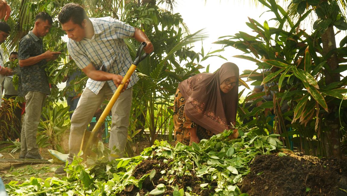 Photograph showing two people engaged in gardening activities in a lush, green outdoor setting. One man uses a hoe to turn soil covered with leaves, while a woman wearing a brown hijab tends to plants nearby.