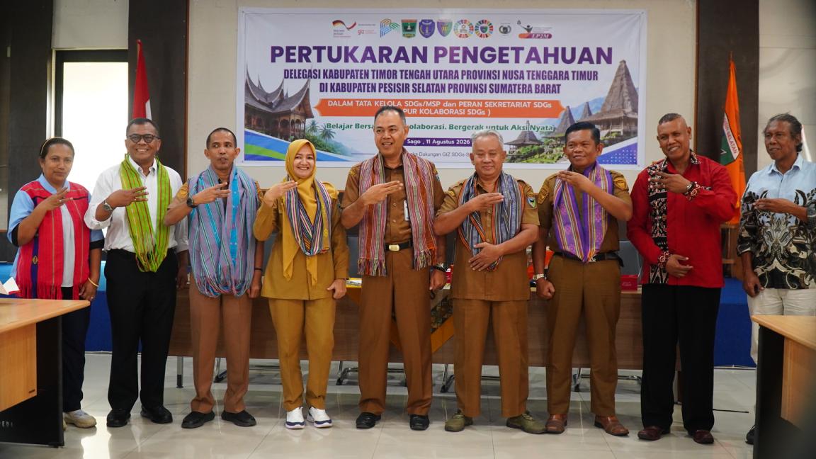 A group of nine people standing in a row indoors, posing for a formal event with a banner behind them titled "Pertukaran Pengetahuan". Participants wear traditional woven scarves and formal attire, with some giving three-fingers gestures, highlighting cultural collaboration and official partnership.