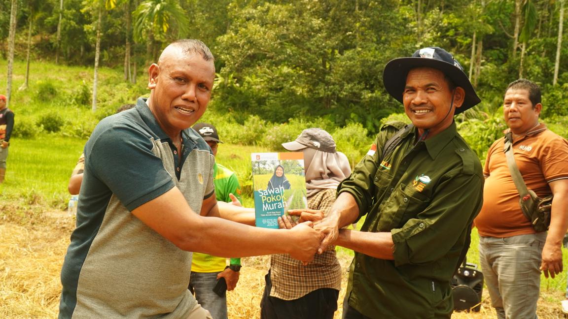 Photograph of two men outdoors shaking hands and holding a book titled "Sawah Pokok Murah" in a rural setting with greenery and other people in the background. The image captures a moment of exchange or presentation, highlighting camaraderie and community engagement.