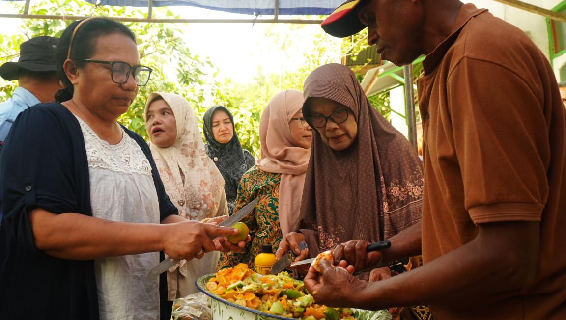 Photograph of a group of people cutting foods outdoors under a canopy. Several individuals, including women wearing headscarves and a man in a brown shirt, are focused on cutting and handling a large bowl of mixed fruit or vegetables.