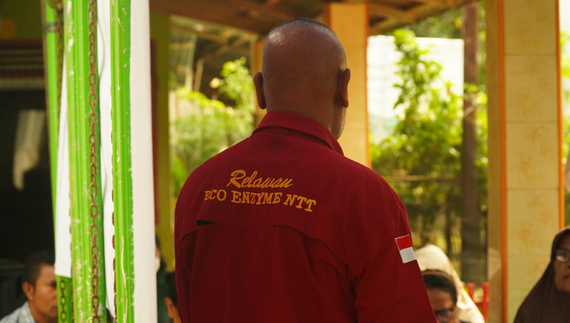 Photograph showing a person wearing a red jacket with yellow text "Relawan ECO ENZYME NTT" and an Indonesian flag patch on the sleeve, standing with their back to the camera.