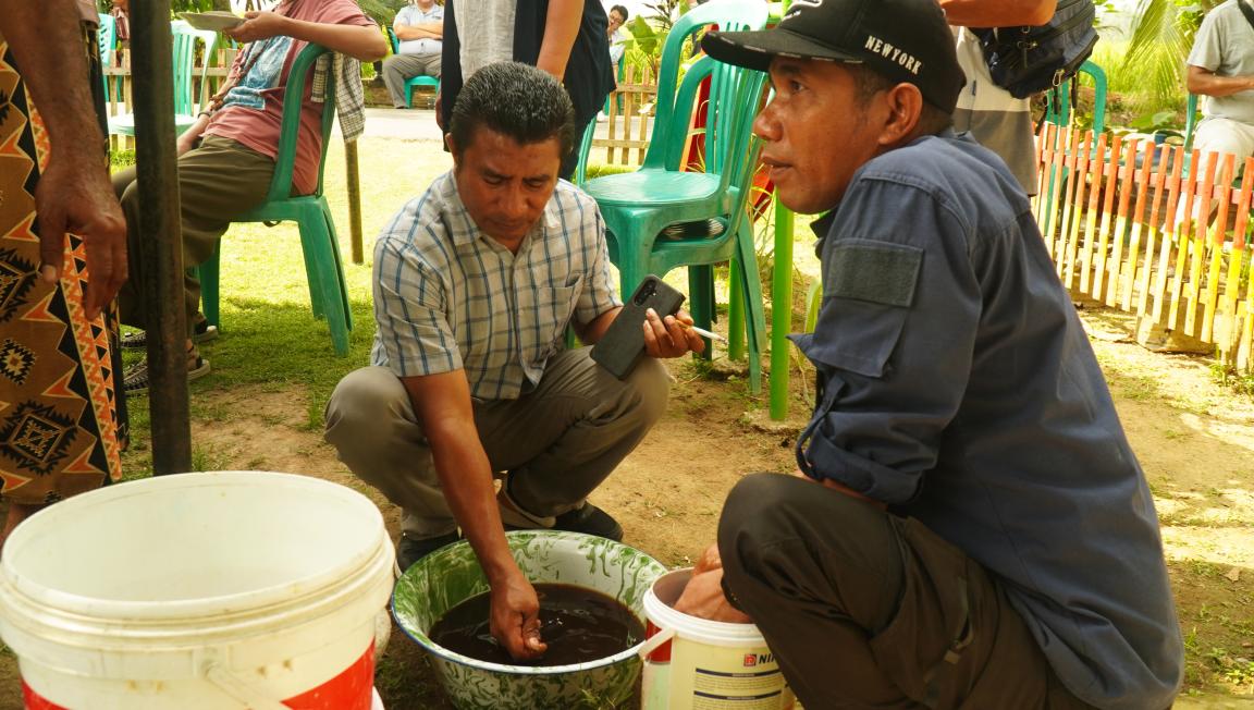 Photograph showing two men crouched outdoors, one stirring a dark liquid in a large metal bowl while the other observes closely. Plastic chairs and a wooden fence are visible in the background, suggesting a communal or workshop setting.