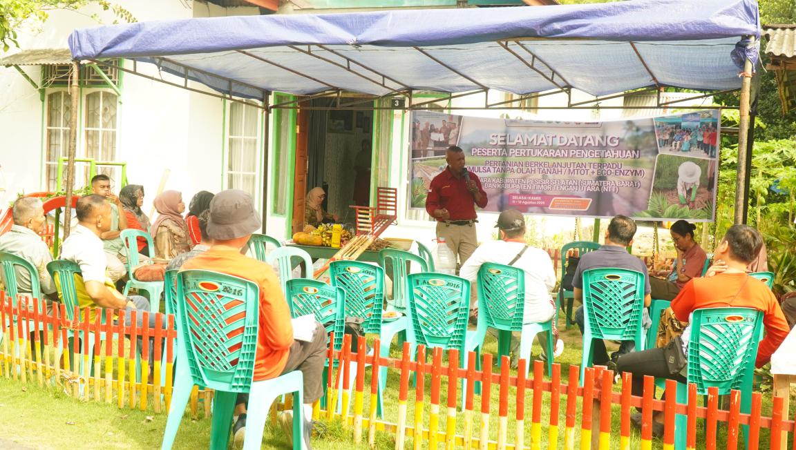 Photograph of an outdoor community meeting under a blue canopy with attendees seated on green plastic chairs facing a speaker. A large banner behind the speaker displays text and images related to agricultural or environmental topics, with a small red and yellow fence enclosing the area.