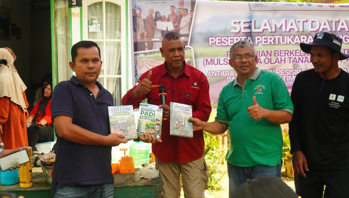 Photograph of four men standing outdoors in front of a banner with Indonesian text, holding and displaying agricultural product packages. The scene highlights a community event focused on sustainable farming practices, with participants showing support and engagement through thumbs-up gestures and product promotion.