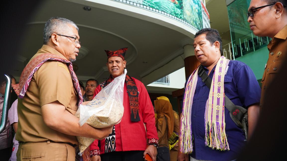 Photograph showing a group of men engaged in a conversation, with one man in a brown uniform holding a clear bag of grains. The setting appears to be a public or official event, with traditional woven scarves worn by some men, indicating cultural or ceremonial significance.