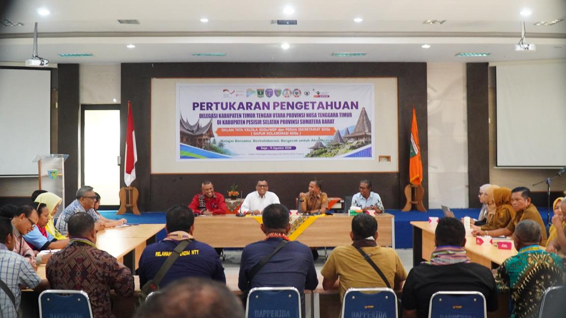 Photograph of a formal meeting or seminar with participants seated around a U-shaped table in a conference room. A banner behind speakers displays Indonesian text about knowledge exchange between regions, with flags and official logos indicating government or organizational involvement.