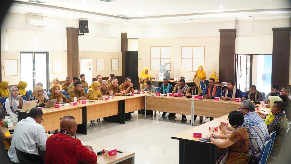 Photograph of a large group meeting in a U-shaped conference room with participants seated around tables, engaging in discussion.