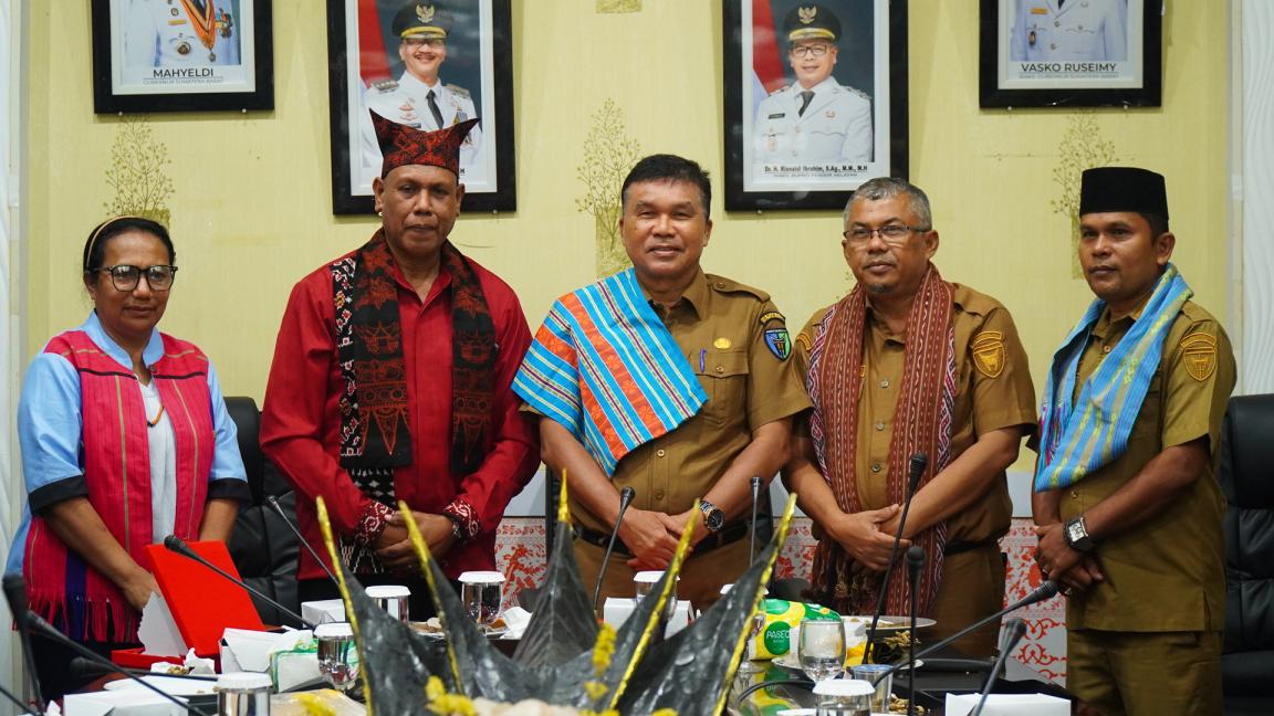 Photograph of five people standing behind a conference table in an office setting, wearing traditional woven sashes over their clothing.