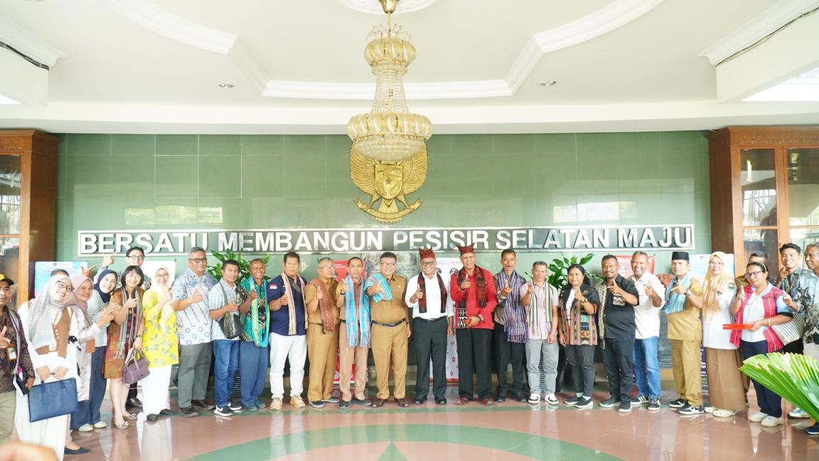 Photograph of a large group of people standing in a semicircle inside a building with a green wall and a chandelier overhead. The group poses in front of a sign reading "BERSATU MEMBANGUN PESISIR SELATAN MAJU," indicating a community or organizational gathering focused on development in South Coastal region.