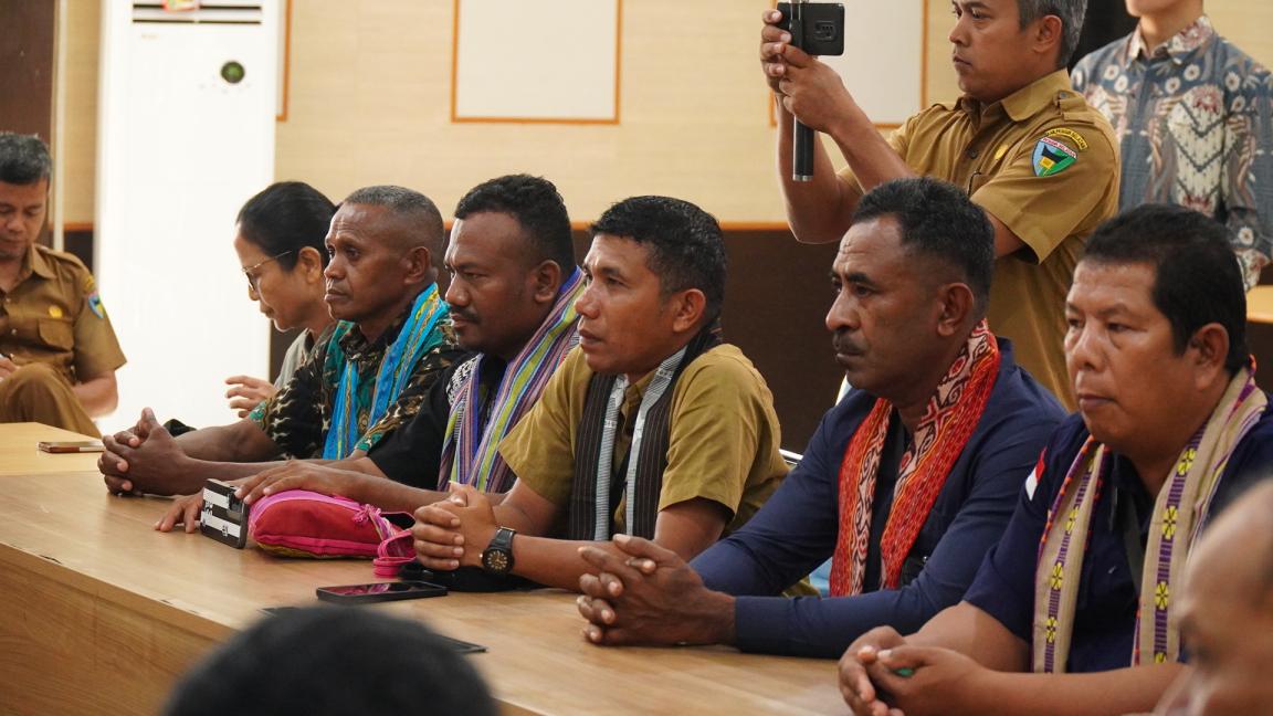Photograph of a group of men seated at a conference table, attentively listening during a formal meeting. Several men wear traditional woven scarves over their clothing, and one person in the background is recording the event with a smartphone.