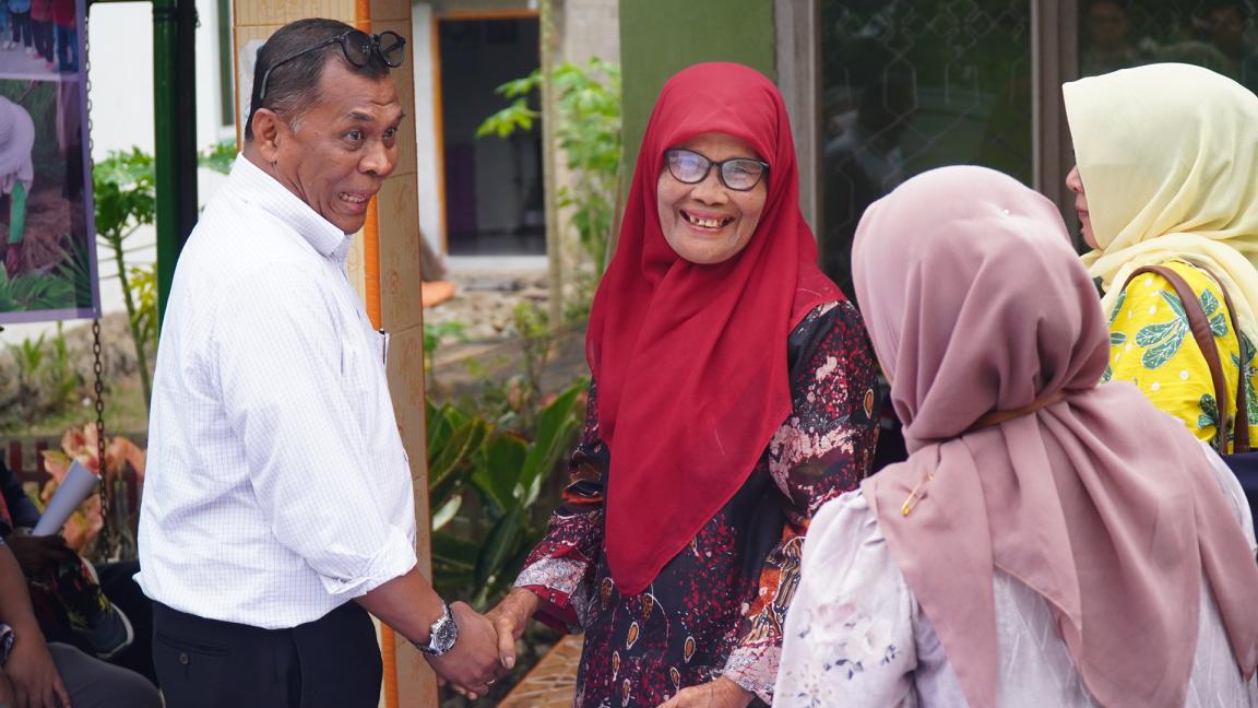 Photograph showing a group of four adults engaged in a friendly outdoor conversation. Two individuals, a man in a white shirt and a woman in a red hijab, are shaking hands and smiling, while two other women wearing hijabs stand nearby.