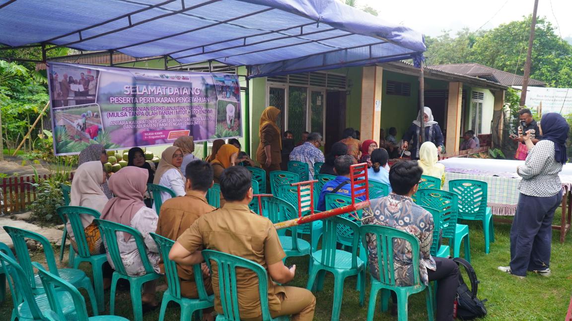 Photograph of an outdoor community meeting or workshop with attendees seated on green plastic chairs under a blue canopy.