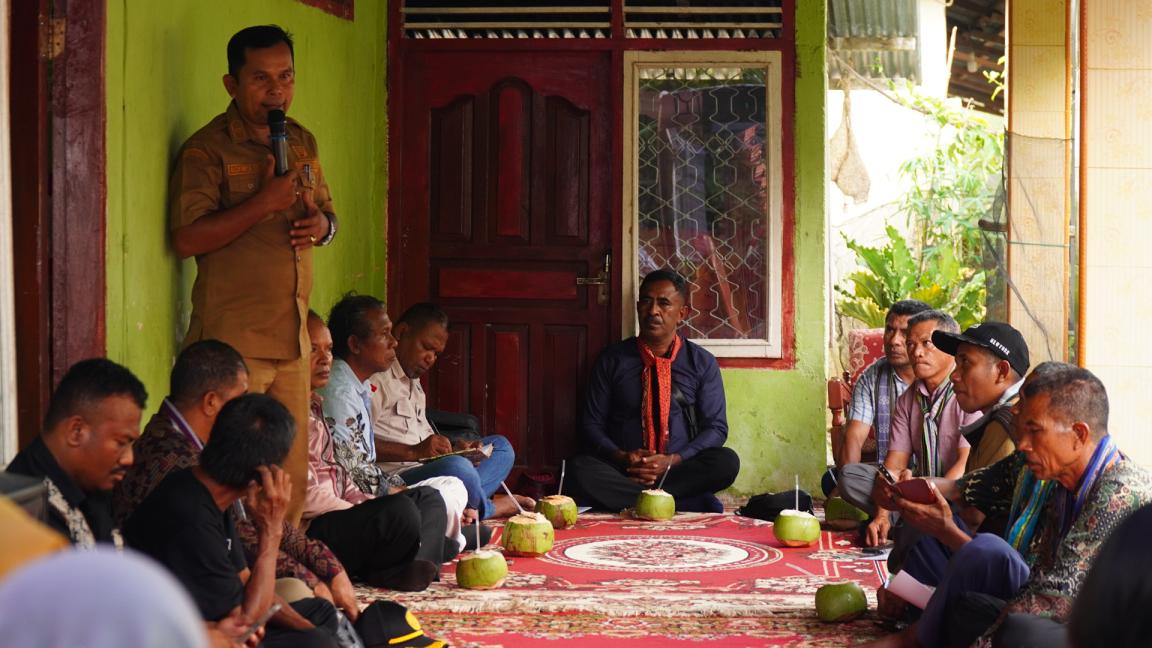Photograph of a group meeting held indoors with men seated on the floor around a red patterned carpet, each with a green coconut drink. One man stands speaking into a microphone, suggesting a formal discussion or community gathering in a modest room with green walls and wooden door and window frames.