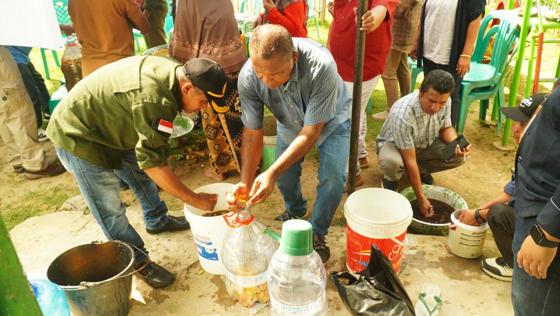 Photograph showing a group of people engaged in a hands-on activity involving water and containers outdoors. Several individuals are filling and handling buckets and bottles, with green plastic chairs and grass visible in the background, suggesting a community or educational event.