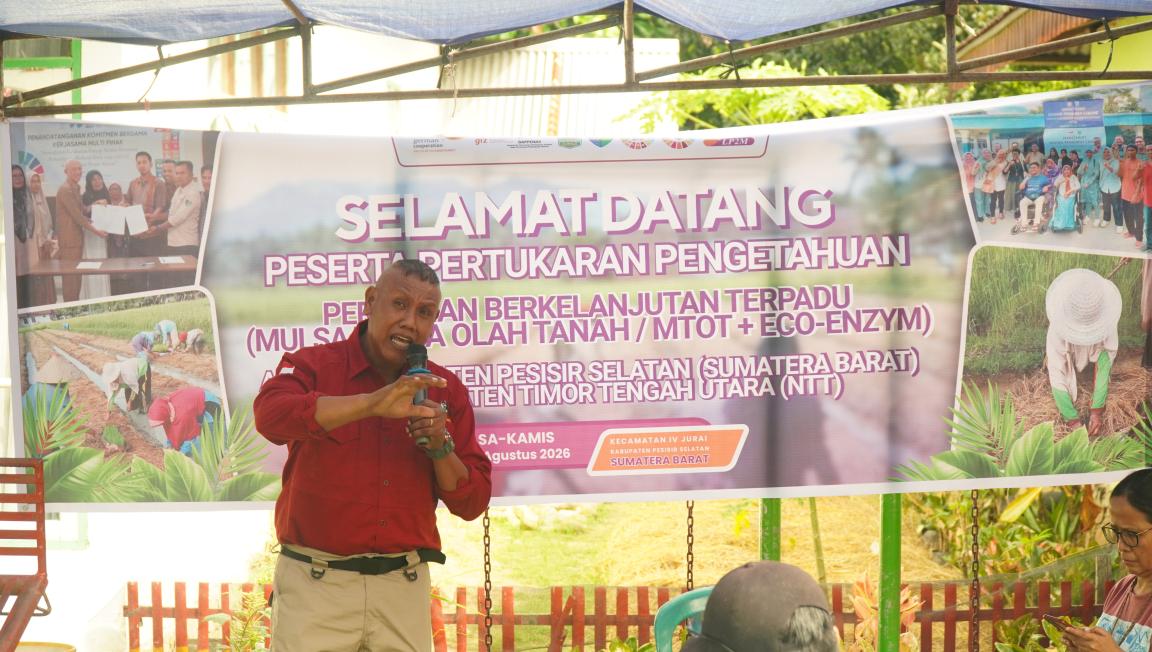 Photograph of a man speaking into a microphone at an outdoor event under a tent, with a large banner behind him. Banner text welcomes participants to a knowledge exchange on sustainable integrated farming techniques in soil processing.