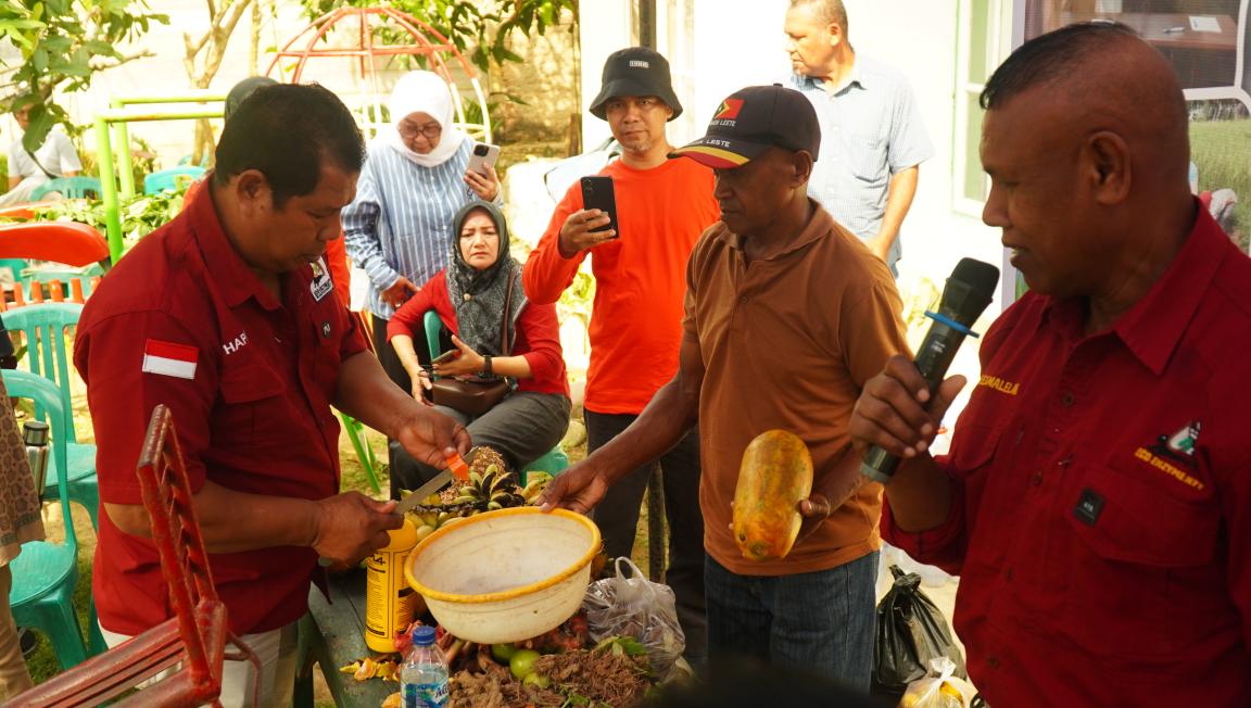 Photograph showing a group of people gathered around a table where a man in a red shirt is preparing fruit using a tool. One man holds a microphone and another holds a yellow fruit, while others observe and record the activity, suggesting a demonstration or educational event.