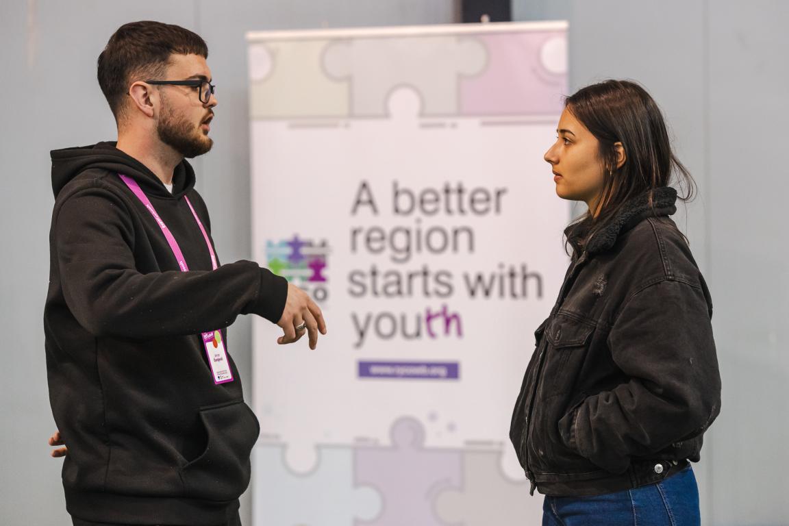 Two young participants talking during Superschools exchange activity, with a RYCO banner reading "A better region starts with youth" in the background.