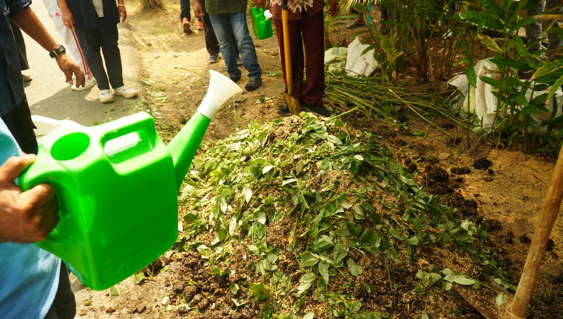 Photograph showing a group of people gathered around a compost pile covered with green leaves and organic material. One person is watering the compost with a bright green watering can, highlighting a composting or gardening activity.