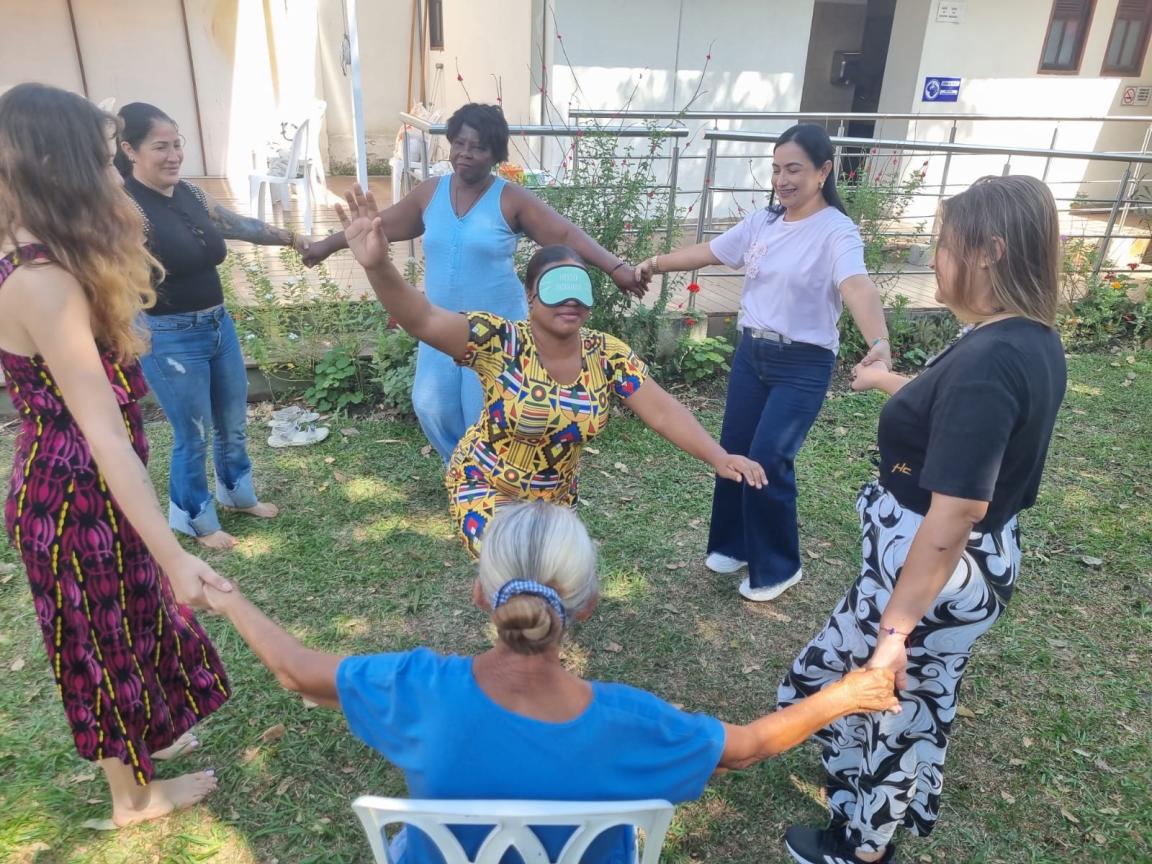Un grupo de mujeres se toman de las manos en un círculo. Una mujer con los ojos vendados está de pie en el medio de círculo. 