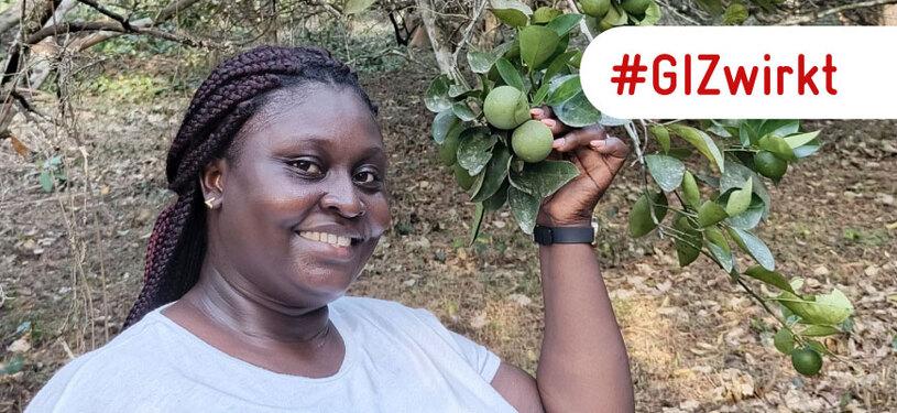 Eine Frau in einem weißen T-Shirt steht lächelnd vor einem Baum. Sie hält einen Zweig mit Limettenfrüchten in der linken Hand. In roter Schrift steht “GIZ wirkt” geschrieben.