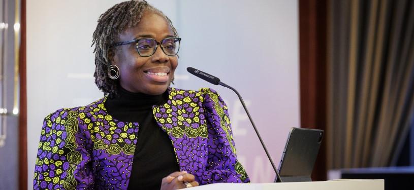 Eine Frau (Mavis Owusu-Gyamfi) in einem lila Blazer steht an einem Podium mit einem Mikrofon und lächelt.