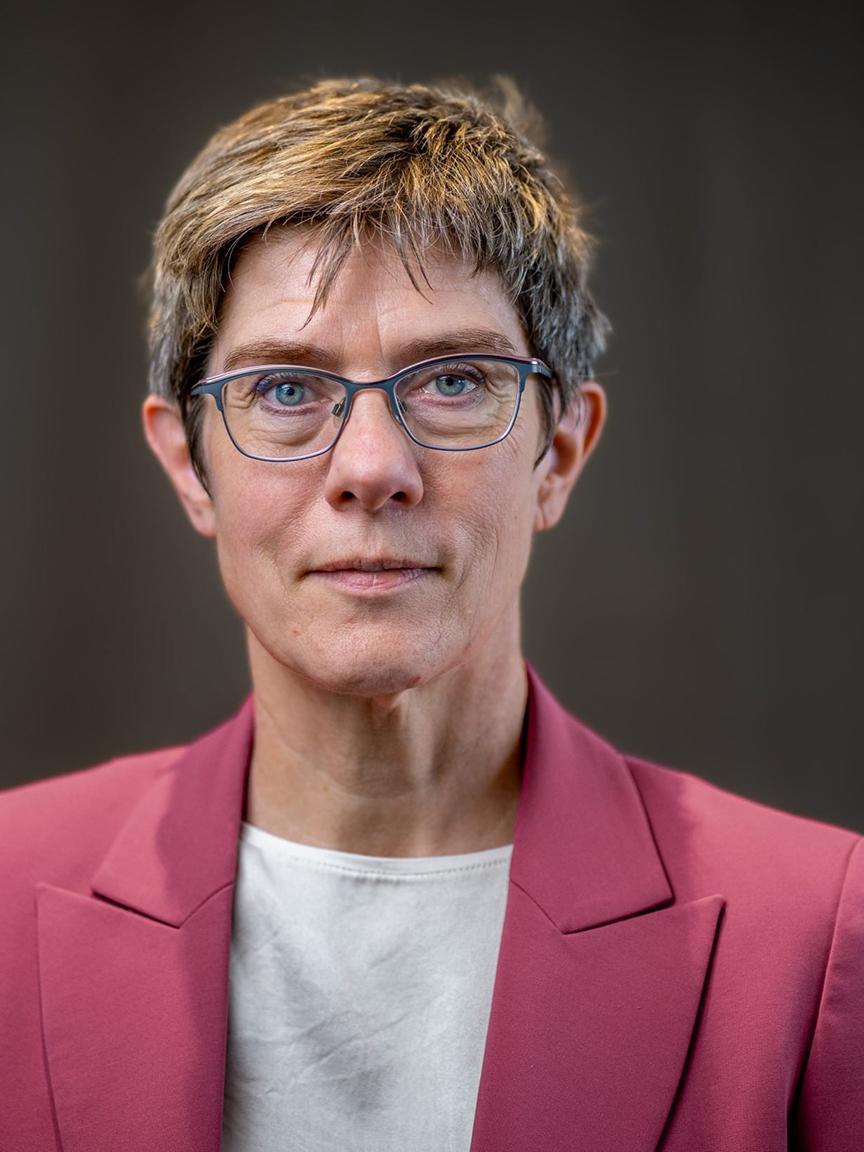 Porträt einer Frau mittleren Alters mit kurzen, gesträhnten Haaren, Brille, weißem Oberteil und rotem Blazer vor neutralem Hintergrund, die direkt in die Kamera blickt.