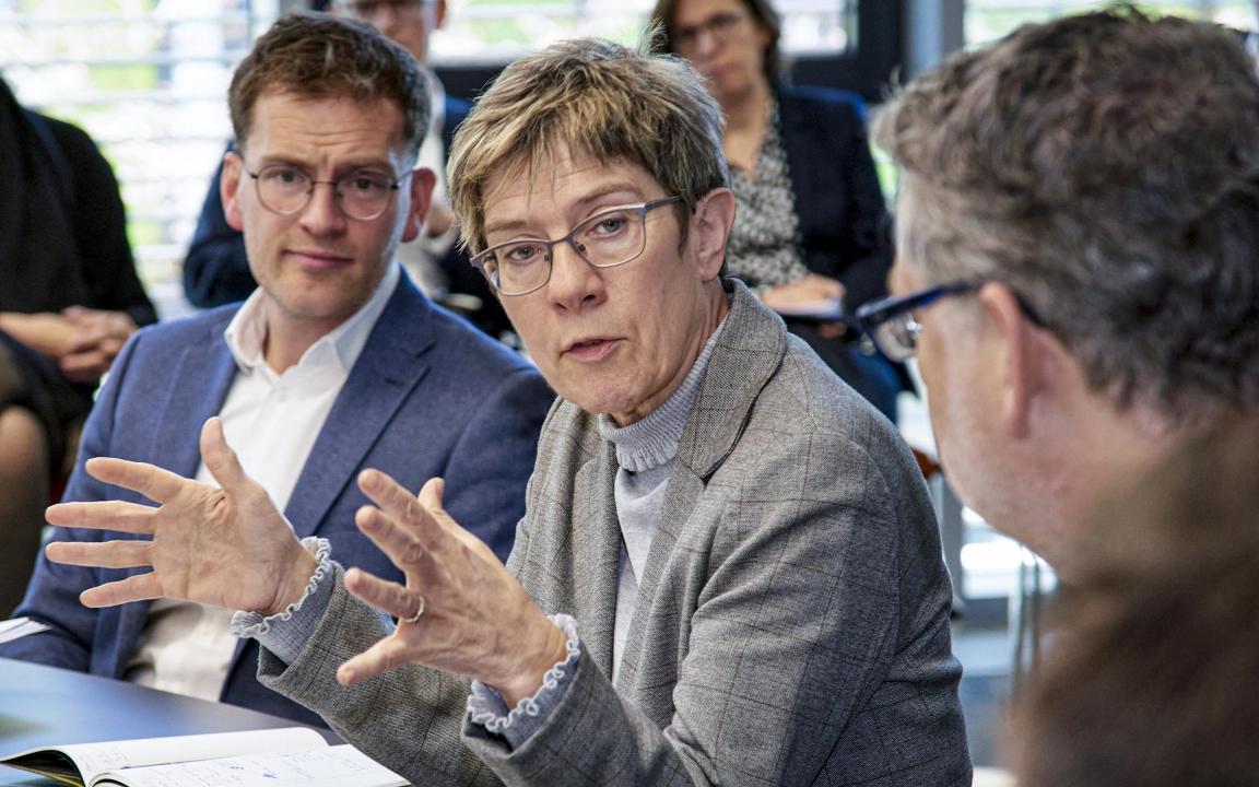 Diskussion in einem Konferenzraum mit einer Frau im grauen Blazer, die mit Gestik spricht, umgeben von mehreren Personen in Businesskleidung, die aufmerksam zuhören.