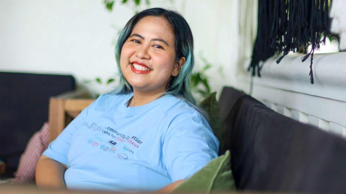 Eine lächelnde Frau mit teilweise türkisfarbenem Haar sitzt auf einem Sofa und trägt ein hellblaues T-Shirt mit Community- und Klimaschutz-Logos.