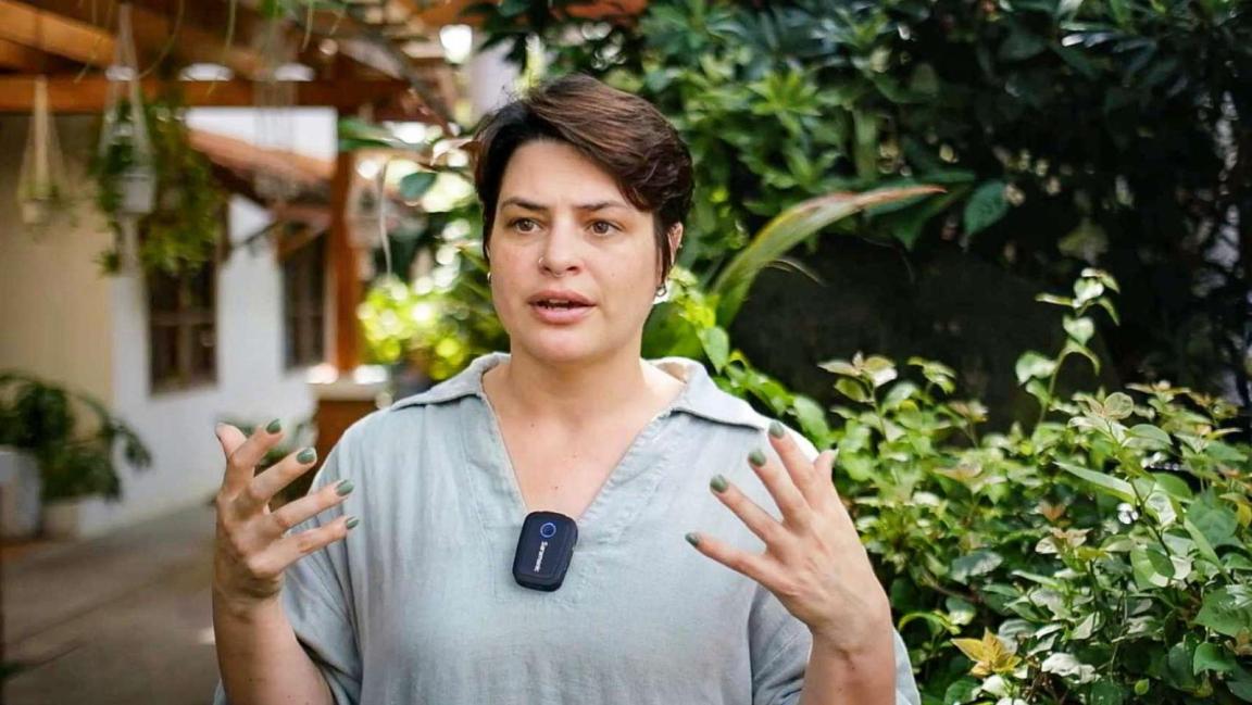 Eine Frau mit Kurzhaarschnitt spricht gestikulierend vor tropischem Grün und trägt ein Mikrofon am Hemd.