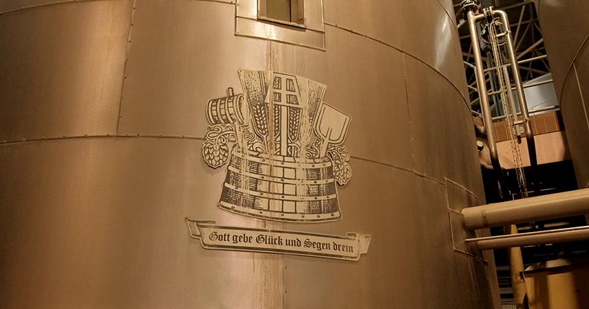 Ein graviertes Emblem auf einer Holzoberfläche, das traditionelle Brauereisymbole darstellt, mit einer Bierkrone in der Mitte, flankiert von Gerstenähren und möglicherweise Brauereiwerkzeugen. Unter dem Emblem befindet sich ein Banner mit der Aufschrift Gott gebe Glück und Segen drein.