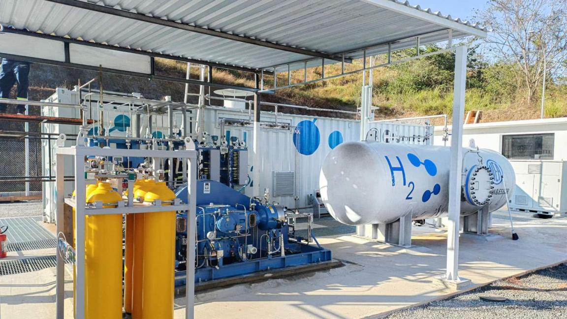 Eine Wasserstoff-Tankanlage mit grauem H₂-Tank und gelben Gasflaschen, überdacht und von technischen Geräten umgeben.