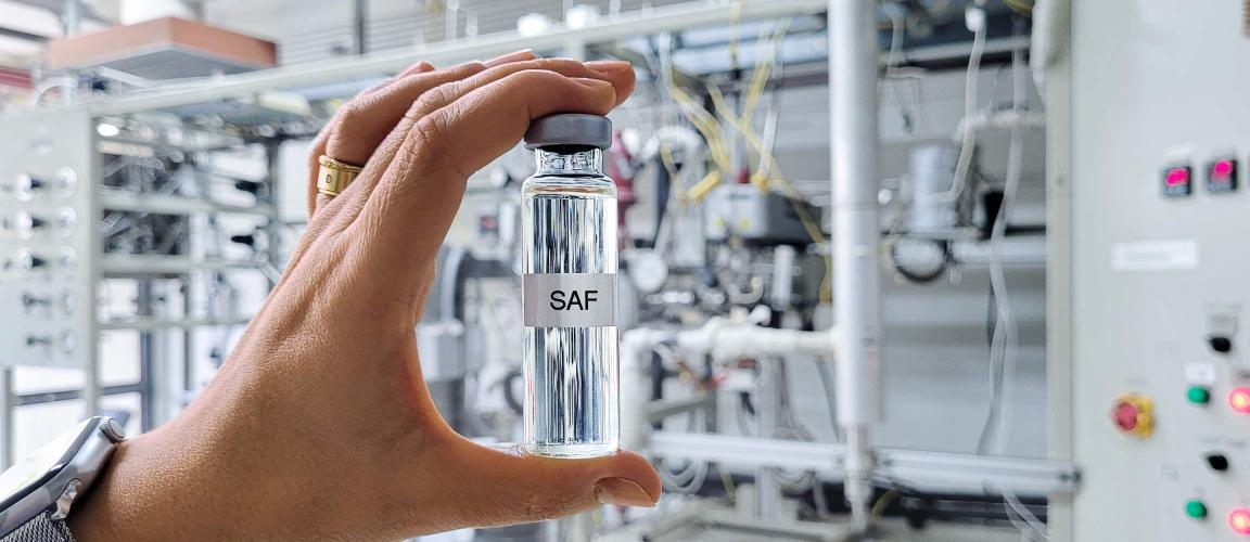 Eine Hand hält ein kleines, mit "SAF" beschriftetes Fläschchen mit klarer Flüssigkeit vor einem verschwommenen industriellen Hintergrund.