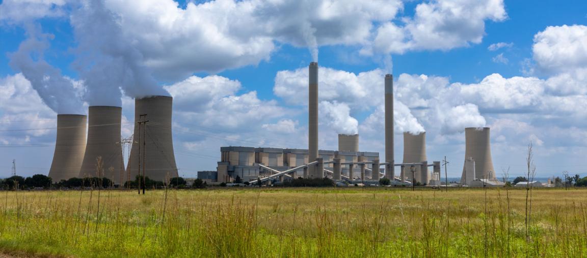 Ein weites Feld im Vordergrund mit einem Kraftwerk im Hintergrund unter einem teilweise bewölkten Himmel. Das Kraftwerk hat mehrere hohe, schlanke Schornsteine und Kühltürme, aus denen Dampf aufsteigt