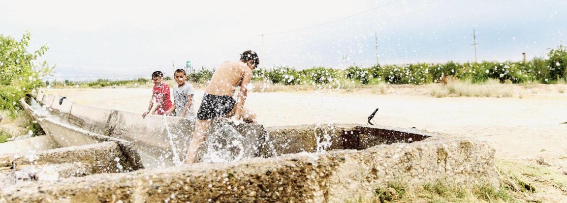 Drei Kinder spielen im Wasser bei sonnigem Wetter. Das vordere Kind ist in Badekleidung und spritzt aktiv Wasser, während die beiden anderen Kinder, eines in Pink und das andere in grau, zuschauen. Sie befinden sich an einem Wasserkanal in Zentralasien.