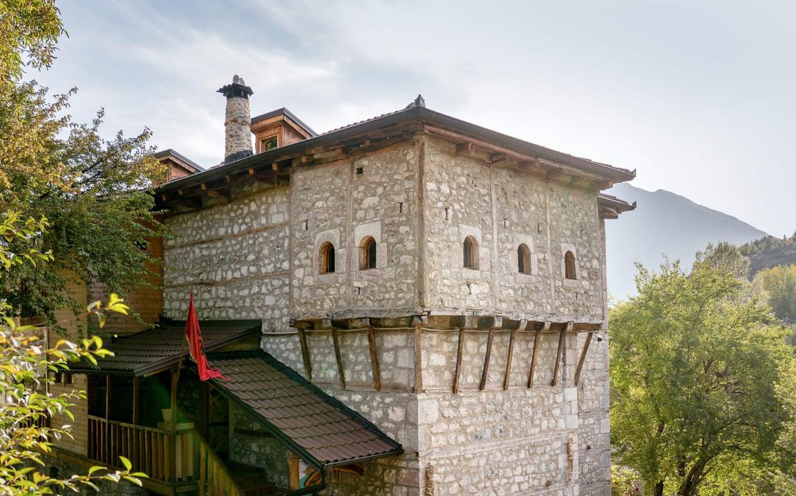 Ein traditionelles Steinhaus mit kleinen Fenstern, Holzdetails und der roten Fahne Albaniens steht inmitten grüner Bäume mit Bergen im Hintergrund.