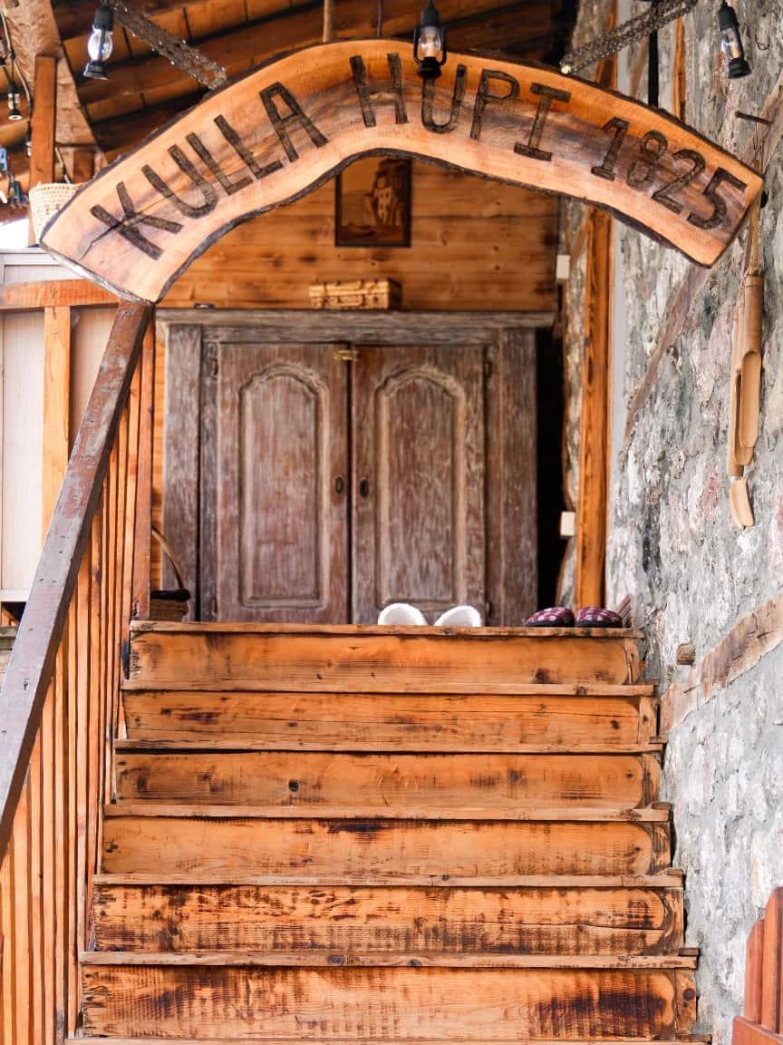 Eine hölzerne Treppe führt zu einem rustikalen Eingang mit einem Holzschild, auf dem „KULA HUPI 1825“ steht.