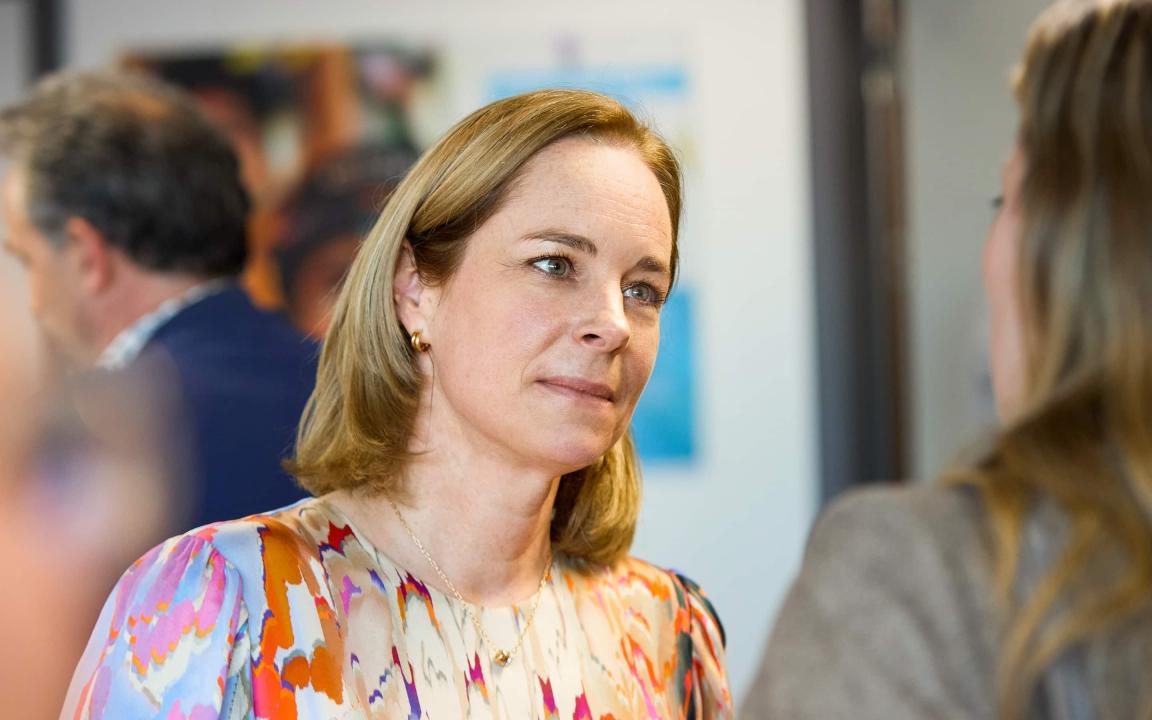 Eine Frau mit schulterlangem blondem Haar und bunter Bluse im Gespräch mit einer anderen Person, im Hintergrund unscharfe Menschen und Bilder an der Wand.