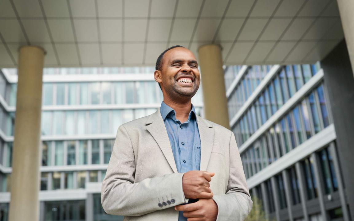 A smiling man in a light-coloured jacket and blue shirt stands in front of a modern office building with a glass façade.