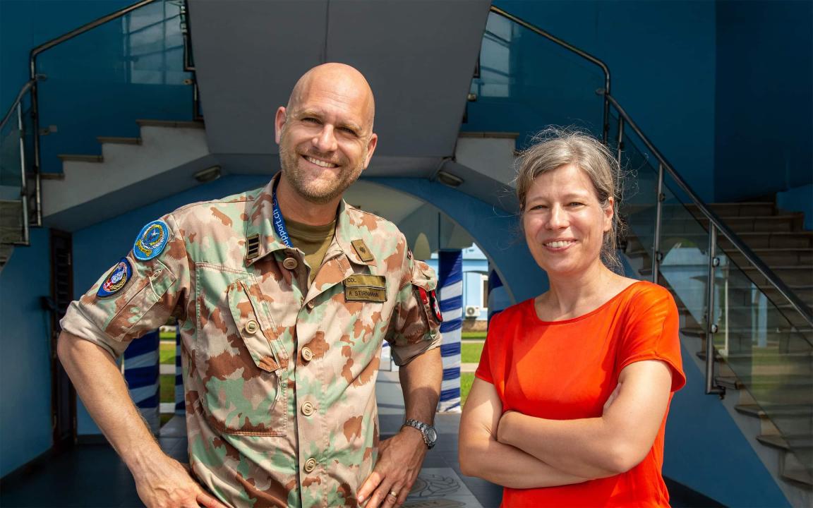 A smiling man in military camouflage uniform with a name tag reading ‘A. Stirnimann’ stands next to a woman in a red shirt in front of a modern blue building.