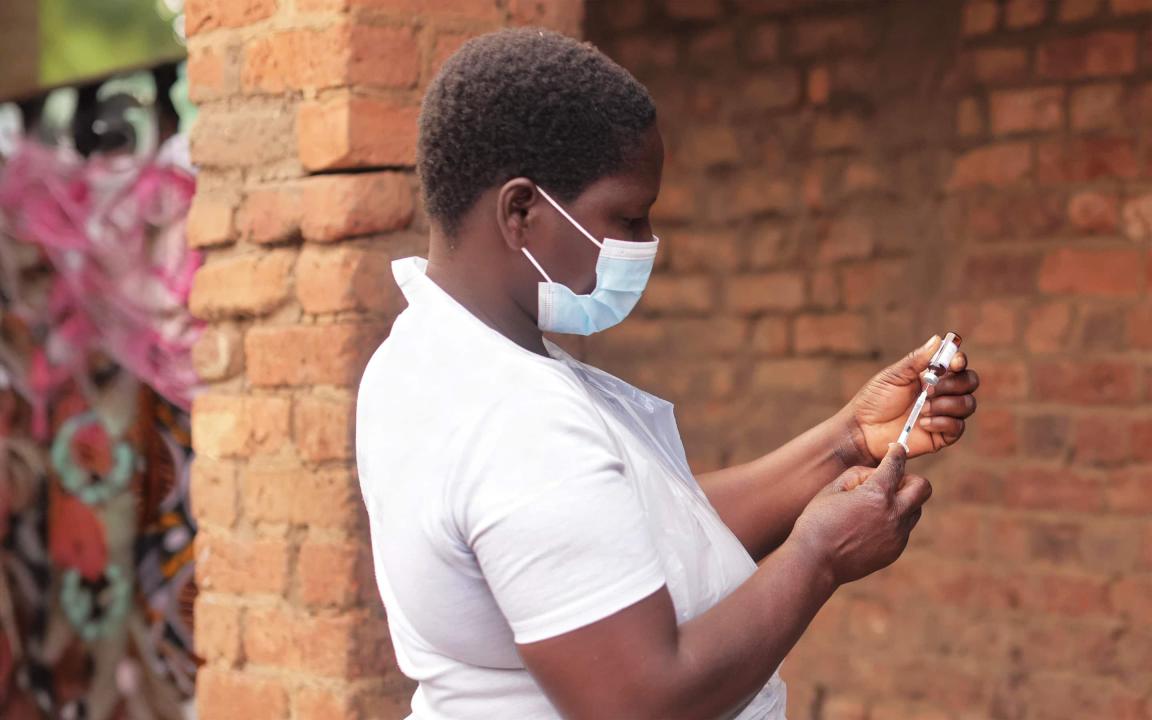 A woman wearing a medical face mask and a white T-shirt draws up an injection from a vaccine vial outdoors.