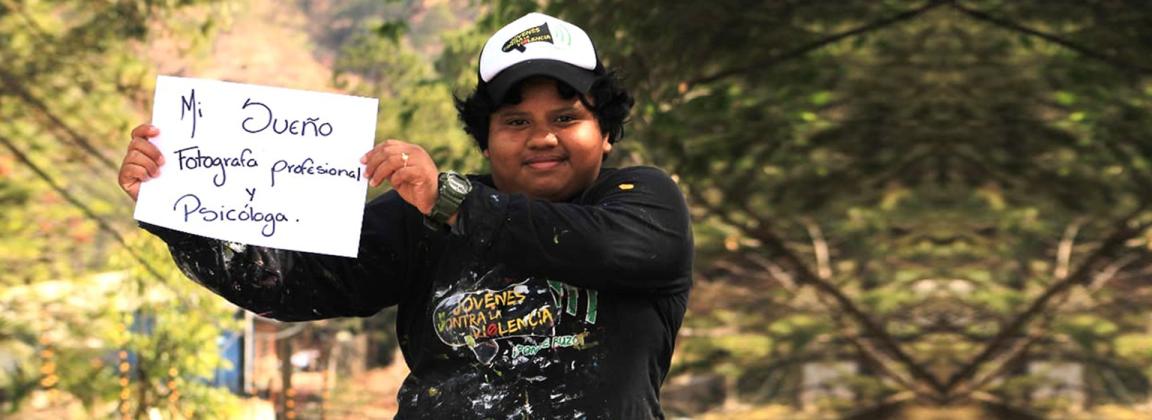 MHPSS Kurs Teilnehmerin.jpeg Eine junge Person mit Schirmmütze und farbbefleckter Kleidung hält einen Zettel hoch. Auf ihm steht: Mi sueño: Fotografa profesional y psychóloga (Mein Traum: Professionelle Fotografin und Psychologin).
