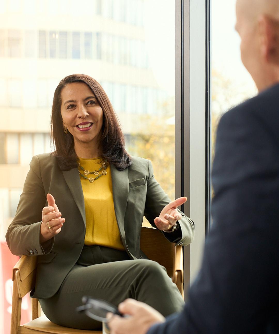 A woman in an olive green suit and yellow blouse is smiling and speaking during a conversation by a window.