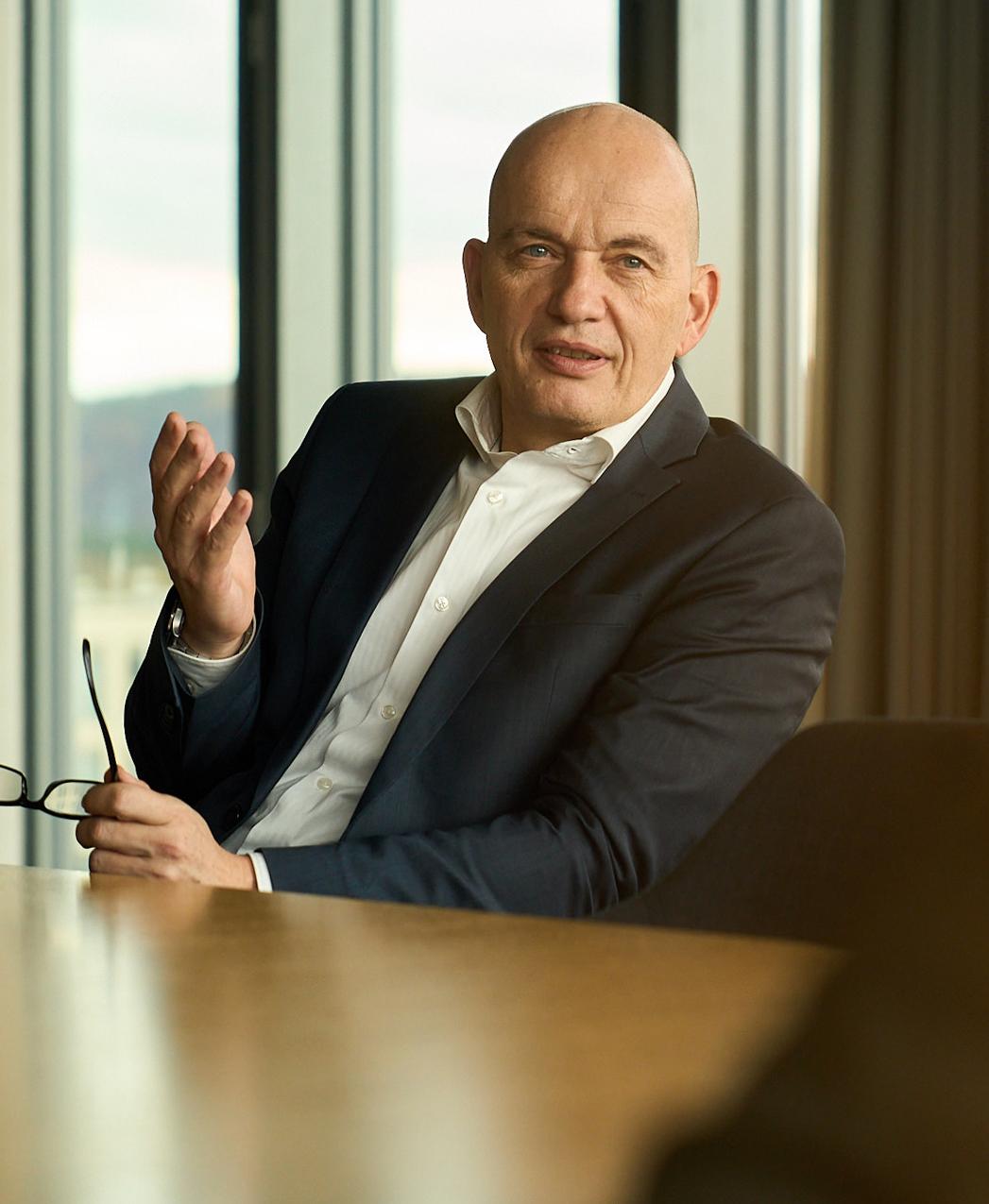 A man in a dark suit holding glasses is speaking in a modern conference room with large windows.