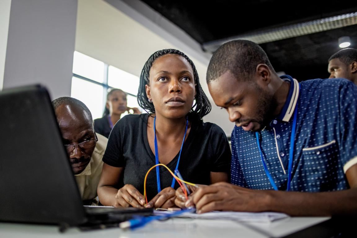 Eine Gruppe von drei Menschen arbeitet mit Kabeln und einem Laptop in einem Vorlesungsraum. Die Frau schaut aufmerksam hoch, die beiden Männer beobachten konzentriert.